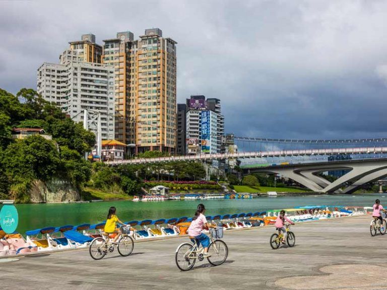 บรรยากาศการพักผ่อนริมน้ำที่มีผู้คนปั่นจักรยานบนทางเท้ากว้างขวาง โดยมีฉากหลังเป็นสะพานแขวนและตึกสูงในย่านเขื่อนซินเตี้ยน ประเทศไต้หวัน