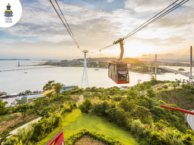 กระเช้าลอยฟ้า 2 ชั้น Queen Cable Car สีแดงขณะเคลื่อนที่ผ่านอ่าวฮาลอง มองเห็นวิวทิวทัศน์เมืองท่าและหมู่เกาะหินปูนจากมุมสูงในช่วงพระอาทิตย์ตกดิน