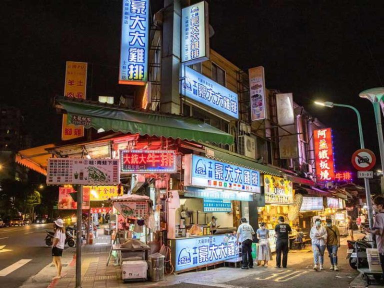 บรรยากาศร้านอาหารแผงลอยในตลาดกลางคืนซื่อหลินที่มีป้ายไฟนีออนสว่างไสวและร้านไก่ทอดเจ้าดัง