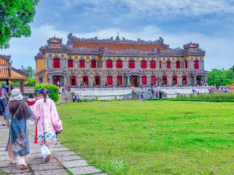 นักท่องเที่ยวสวมชุดอ่าวหญ่ายเดินชมความงามของสถาปัตยกรรมโบราณของพระราชวังเมืองเว้ที่มีหน้าต่างสีแดงโดดเด่นและลวดลายแกะสลักสวยงาม