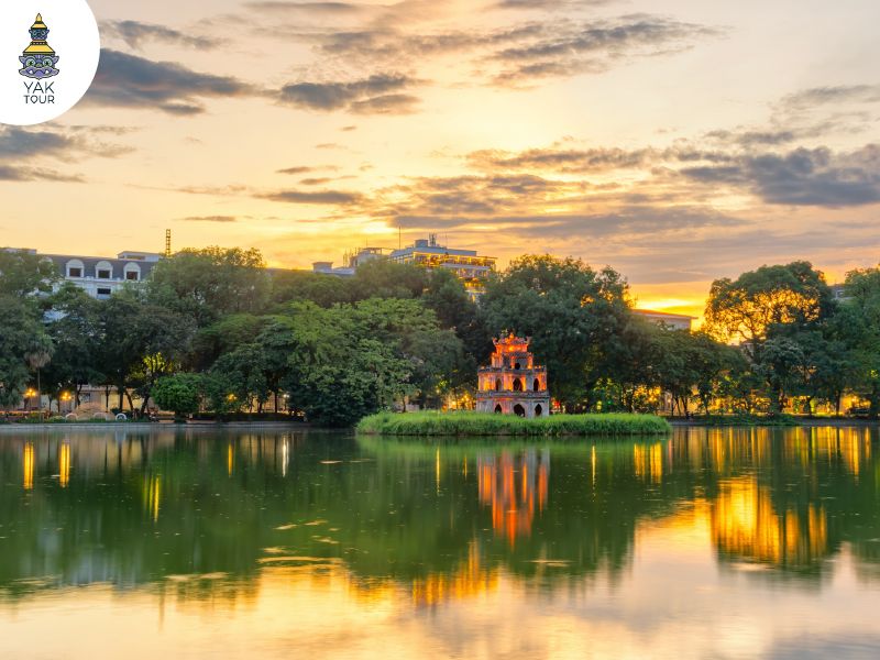 ทัศนียภาพยามเย็นของทะเลสาบคืนดาบ (Hoan Kiem Lake) พร้อมแสงไฟระยิบระยับจากหอคอยเต่าที่สะท้อนบนผิวน้ำ รายล้อมด้วยพื้นที่สีเขียวใจกลางฮานอย