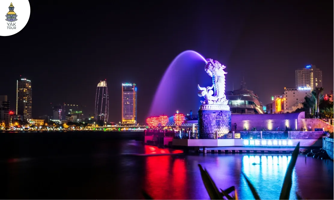 บรรยากาศยามค่ำคืนที่คาร์ฟดราก้อน (Ca Chep Hoa Rong) ริมแม่น้ำฮัน แสดงรูปปั้นปลาคาร์ฟหัวมังกรพ่นน้ำเรืองแสง พร้อมทิวทัศน์เมืองดานังที่เต็มไปด้วยแสงไฟสวยงาม