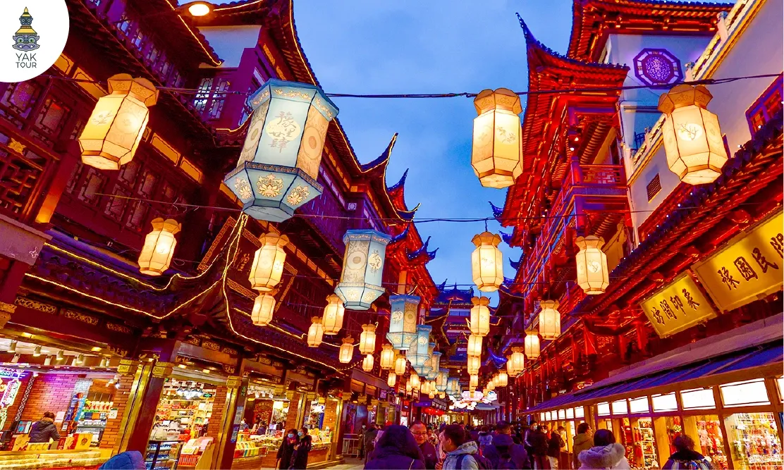 บรรยากาศยามเย็นที่ตลาดร้อยปีเฉิงหวังเมี่ยว (Chen Wang Miao) เซี่ยงไฮ้ รายล้อมด้วยอาคารสถาปัตยกรรมจีนโบราณสีแดง ตกแต่งด้วยโคมไฟจีนโบราณและโคมไฟสีฟ้าสว่างไสว มีนักท่องเที่ยวเดินชมร้านค้าและสตรีทฟู้ด