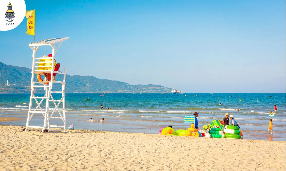 ชายหาดไชน่า (China Beach) หรือหาดหมีเคว ดานัง เห็นหอคอยไลฟ์การ์ดสีขาวบนพื้นทราย มีห่วงยางหลากสีวางเรียงราย และวิวทะเลสีครามที่มีฉากหลังเป็นภูเขาเขียวขจี