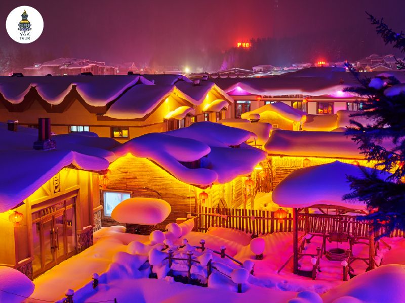 บรรยากาศยามค่ำคืนของหมู่บ้านหิมะที่มีหิมะหนานุ่มปกคลุมหลังคาบ้านไม้คล้ายรูปเห็ด ตกแต่งด้วยโคมไฟสีแดงและแสงไฟสีอุ่นทั่วทั้งบริเวณ