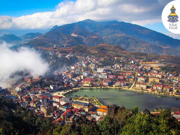 ทิวทัศน์มุมสูงของเมืองซาปาจากยอดเขาฮามรอง มองเห็นบ้านเรือนหลากสีสันรายล้อมทะเลสาบซาปาและมีฉากหลังเป็นเทือกเขาสูงท่ามกลางไอหมอก