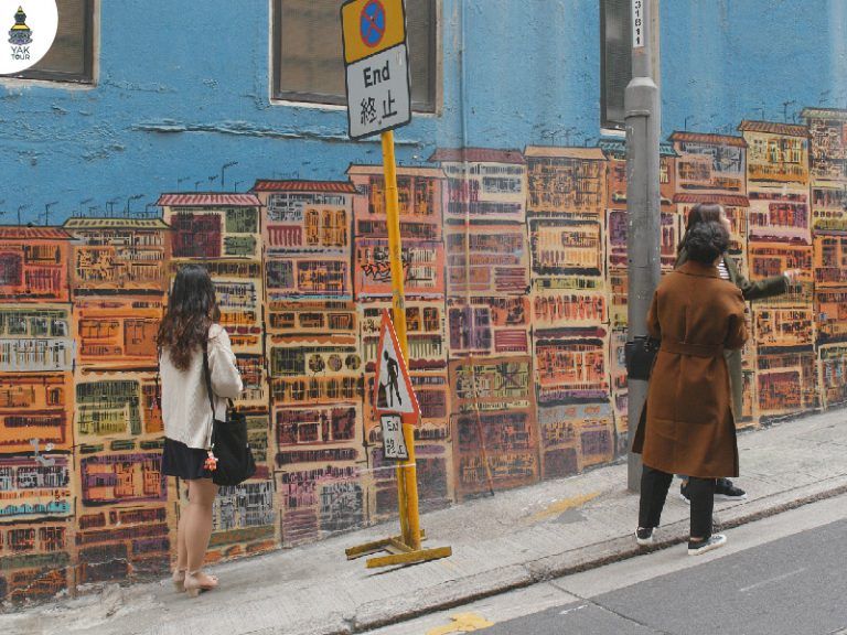 นักท่องเที่ยวกำลังยืนชมภาพวาดสตรีทอาร์ทรูปตึกแถวหลากสีสันบนกำแพงสีฟ้าสดใส ซึ่งเป็นจุดเช็กอินยอดฮิตของ ที่เที่ยวฮ่องกง บริเวณริมถนนฮอลลีวู้ด