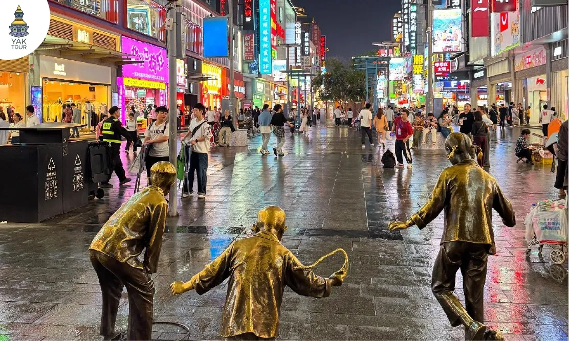 รูปปั้นเด็กทองเหลืองโบราณบนถนนคนเดินหวงซิงลู่ (Huangxing Walking Street) ท่ามกลางแสงสีของป้ายไฟร้านค้าในยามค่ำคืน