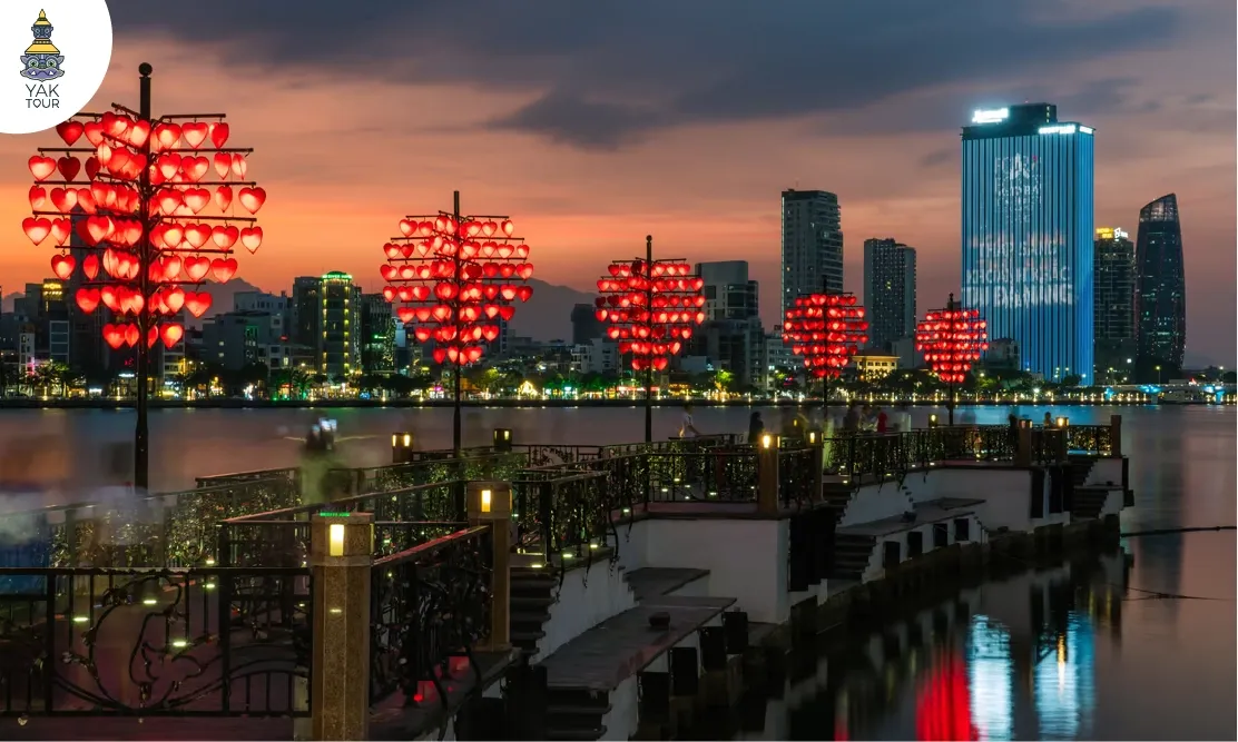 บรรยากาศยามค่ำคืนที่สะพานแห่งความรักดานัง (Love Bridge Da Nang) ริมแม่น้ำฮัน แสดงเสาโคมไฟรูปหัวใจสีแดงส่องสว่างเรียงรายตลอดแนวสะพาน พร้อมทิวทัศน์ตึกสูงเมืองดานังในพื้นหลัง
