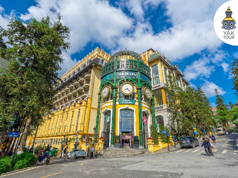 อาคาร Sun Plaza Sapa สีเหลืองมัสตาร์ดสไตล์ฝรั่งเศส โดดเด่นด้วยประตูทางเข้าทรงโค้งและหอนาฬิกาขนาดใหญ่ใจกลางเมืองซาปา ภายใต้ท้องฟ้าสดใส