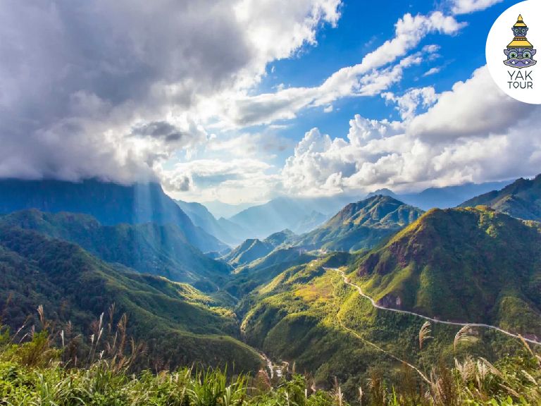 มุมกว้างจากจุดชมวิว Tram Ton Pass มองเห็นเทือกเขาสูงสลับซับซ้อนและเส้นทางถนนคดเคี้ยวพาดผ่านหุบเขา ภายใต้แสงแดดที่ลอดผ่านหมู่เมฆลงมาสู่ยอดเขาในซาปา