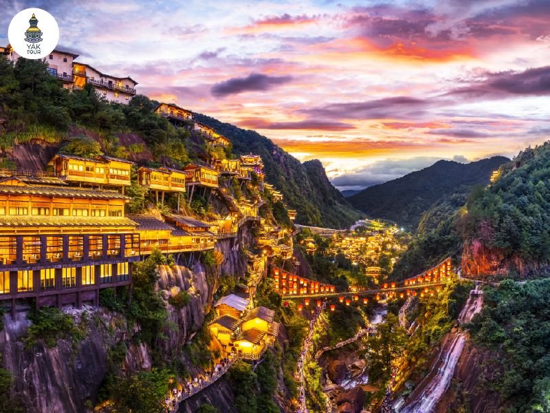 หมู่บ้านโบราณวั้งเซียนกู่บนหน้าผาสูงชันยามเวลาค่ำคืนที่เปิดไฟประดับสว่างไสวท่ามกลางหุบเขาและน้ำตก